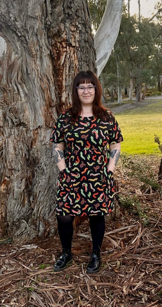 Woman with brown hair, glasses, and arm tattoos wearing black dress with colorful dinosaur pattern, posed against large eucalyptus tree in park